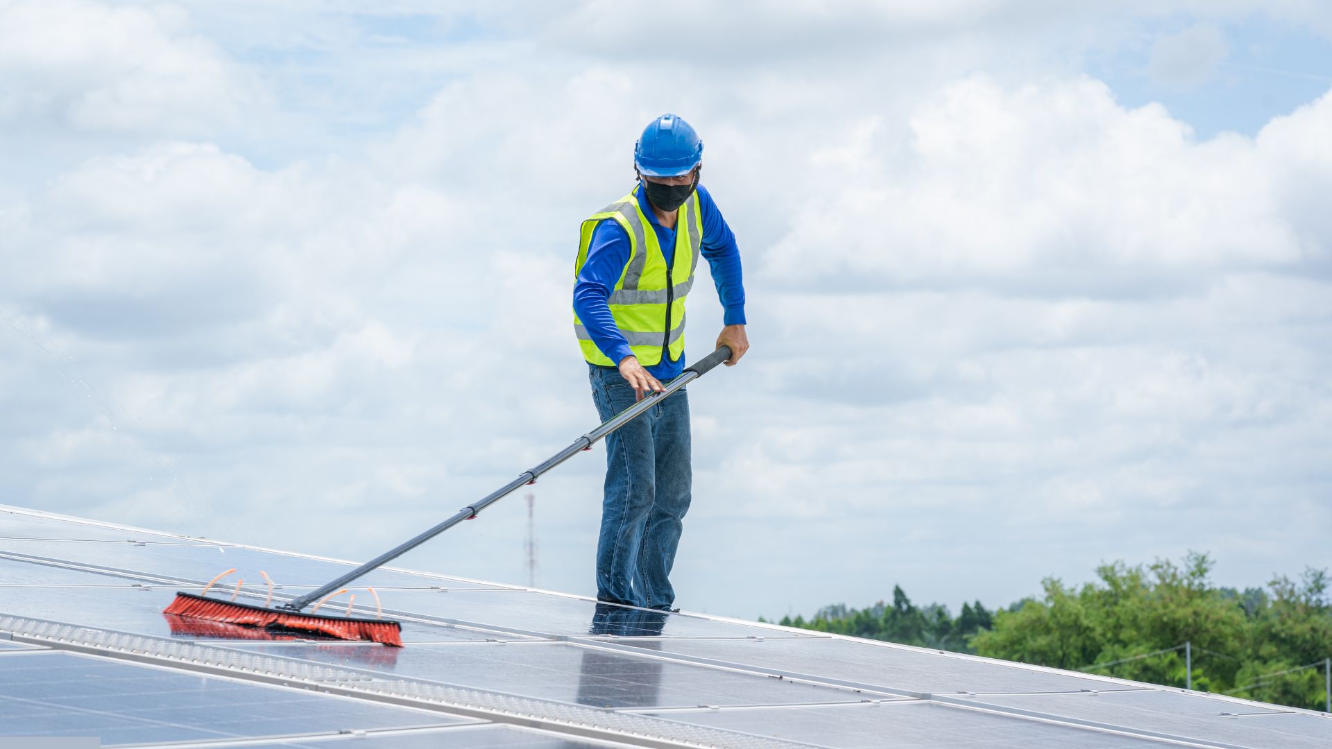 Worker in blue uniform and safety helmet cleaning solar panels using a long-handled brush on a sunny day.