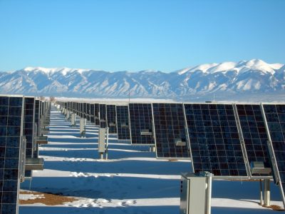 Banks of solar panels in a snowy field by grey mountains, beneath a clear blue sky